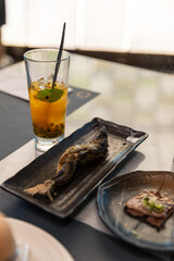 Grilled whole fish served on a black ceramic plate beside a slice of grilled meat and a tall glass of iced passion fruit tea garnished with mint