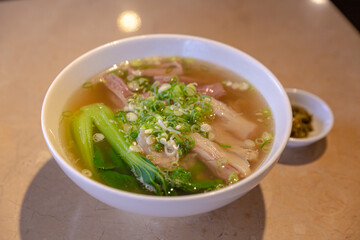 Hot clear beef broth noodle soup with slices of brisket and tendon, fresh bok choy, and green onions served in a ceramic bowl