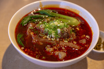 Close-up of spicy beef noodle soup with brisket and tendon in red chili oil broth, garnished with bok choy and scallions in a white ceramic bowl