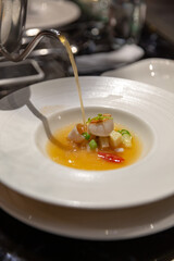 Hot consommé being poured into a dish with scallops and vegetables, a refined presentation of seafood soup in fine dining setting