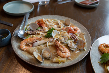 Plate of stir-fried flat noodles topped with assorted seafood including prawns, clams, and squid, served in savory sauce on a wooden table