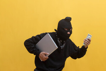 Portrait of a thief dressed in black wearing ski mask balaclava stealing laptop and dollar...