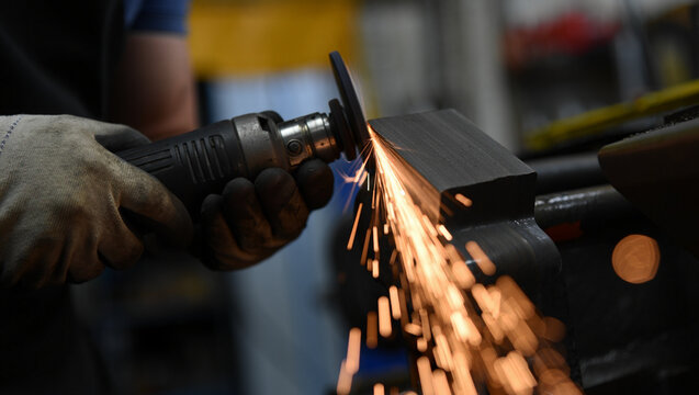 A worker wearing protective gloves operates an electric grinding tool against a metal workpiece, creating a controlled arc of golden sparks