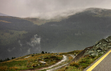 Moody mountain scene with fog and cloudy sky. Wet winding road after rain, caravan in the middle,...