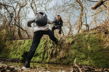 Naklejka premium Two young men help each other navigate a forest trail during a winter hike. Teamwork and friendship are shown against a serene and mossy natural backdrop.