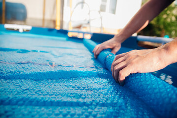 The blue pool cover is assembled into a roll by men's hands, the concept of summer recreation and...