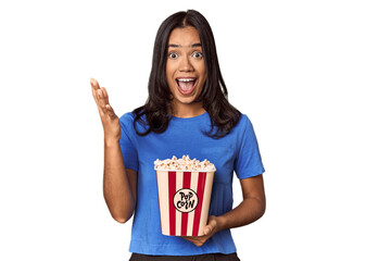 Young woman with popcorn bucket in studio receiving a pleasant surprise, excited and raising hands.