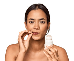 Filipino woman holds deodorant in studio with fingers on lips keeping a secret.
