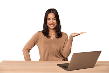 Filipino woman at desk with laptop showing a copy space on a palm and holding another hand on waist.
