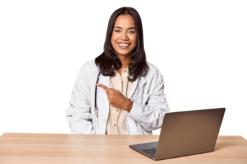 Young Filipino doctor with laptop in studio smiling and pointing aside, showing something at blank space.
