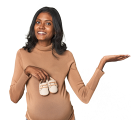 Pregnant Indian with baby shoes showing a copy space on a palm and holding another hand on waist.