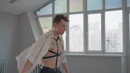 shirtless boy dressed in black pants raises arms to put on white shirt in bright modern room with large window panels, natural daylight highlighting upper body against urban cityscape backdrop