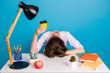 Tired student resting at desk holding cup of coffee surrounded by study supplies against a vibrant blue background