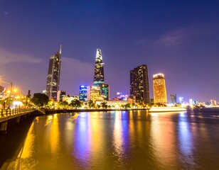 Obraz premium A vibrant cityscape at night, illuminated skyscrapers reflecting on a calm river. The city's modern architecture glows with colorful lights, showcasing a bustling urban scene.