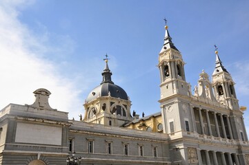 Obraz premium A stunning view of the Almudena Cathedral in Madrid, Spain, showcasing its grand neoclassical facade, bell towers, and central dome against a bright blue sky. 