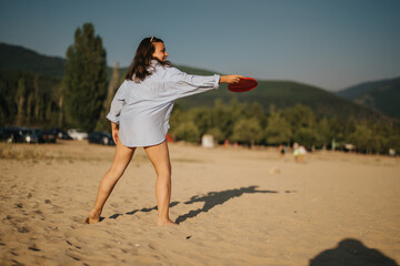 A cheerful woman enjoys a sunny day at the beach, throwing a frisbee. The sandy area, green hills,...