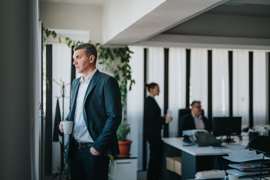 Businessman stands near a window with a coffee while colleagues discuss work behind.