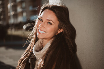 Charming young woman with long brunette hair smiling outdoors in autumn sunlight wearing stylish...