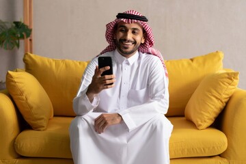 Smiling saudi arab man using smartphone on yellow couch at home modern technology lifestyle