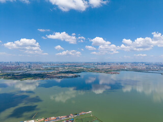 Aerial view of Tangxun Lake reflecting bright blue sky and white clouds, with the sprawling city of...