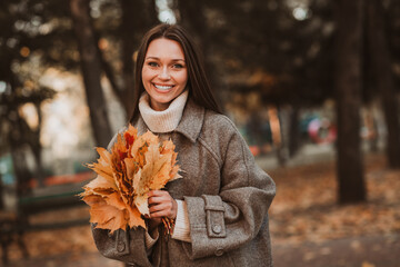 Young woman smiling with colorful autumn leaves in a sunny park during a casual outing, enjoying...