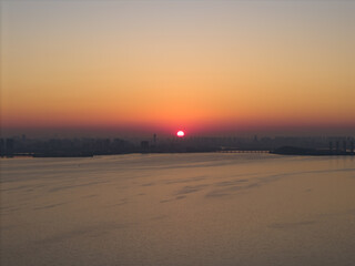 Obraz premium Bright red sun sets gently over Tangxun Lake in Jiangxia District, Wuhan, China, casting beautiful golden light across tranquil water and the distant urban skyline.