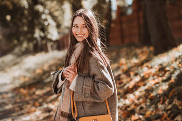 Charming young woman in autumn park enjoying sunshine, wearing stylish coat and scarf, surrounded...