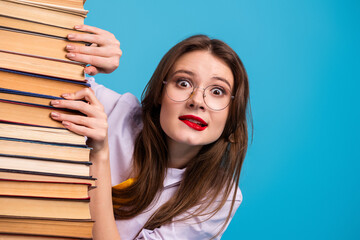 Surprised young student hiding behind a stack of book against a bright blue background, showcasing...