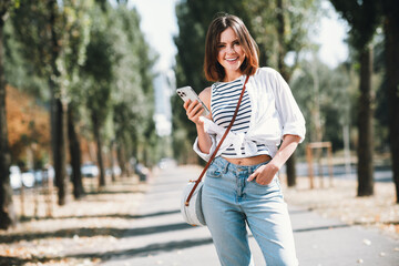Charming young woman in stylish casual outfit enjoying a sunny day outdoors with her smartphone