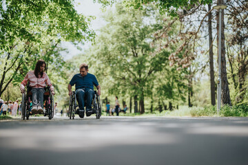 Obraz premium A couple sharing a joyful outing while utilizing wheelchairs in a scenic park environment under the sunny sky and vibrant foliage.