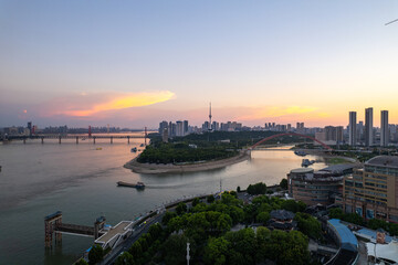 Naklejka premium Wuhan China cityscape offers a stunning panoramic view of the wide Yangtze River at sunset. Bridges span the water, connecting urban areas with many tall buildings under a colorful evening sky.