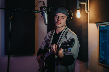 A focused musician wearing headphones plays his guitar while recording in a dimly lit studio. The environment highlights the creativity and dedication involved in music production.