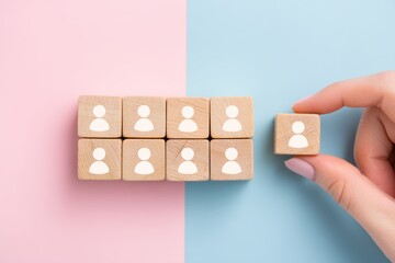 Wooden blocks with white human icons are arranged on a pastel pink and blue background, with a hand picking up one block