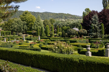 A classic French-style formal garden with intricate geometric hedge patterns, manicured shrubs, and colorful roses, set against a backdrop of rolling hills and lush green trees under a bright blue sky