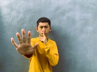 An Asian man in a yellow t-shirt makes a stop gesture with an open palm, indicating a halt or...