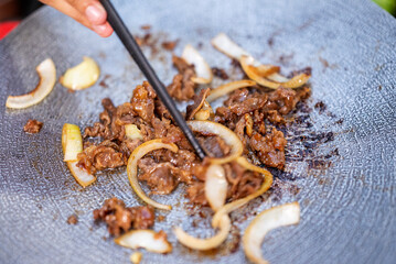 Closeup view of people with chopsticks cooking sliced marinated beef bulgogi on a round grill pan with onions. The scene of the lifestyle of enjoying food in a natural setting