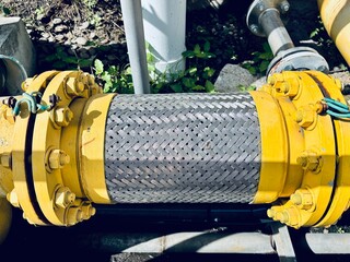 A close-up of a braided stainless steel flexible pipe with yellow flanges and bolts, connecting rigid pipes in an industrial setting.