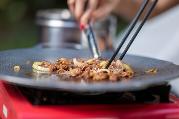 Closeup view of people with chopsticks cooking sliced marinated beef bulgogi on a round grill pan with onions. The scene of the lifestyle of enjoying food in a natural setting