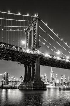 Fototapeta New York City black and white night view of the Manhattan Bridge from DUMBO. Illuminated Lower Manhattan skyline with skyscrapers reflected on the East River