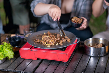 Closeup view of people with tongs cooking sliced marinated beef bulgogi on a round grill pan with onions. The scene of the lifestyle of enjoying food in a natural setting