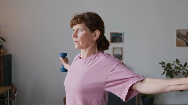 Medium close-up of focused elderly Caucasian woman wth red hair in ponytail doing repeats of bicep curl and chest fly weight training exercise using dumbbells, alone at home