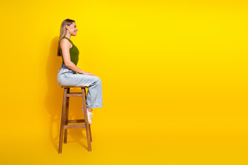 Side view of a cheerful woman in a trendy casual outfit sitting on a stool against a vibrant yellow background