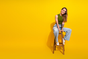 Young woman smiling while sitting on a chair against a vibrant yellow background wearing trendy...