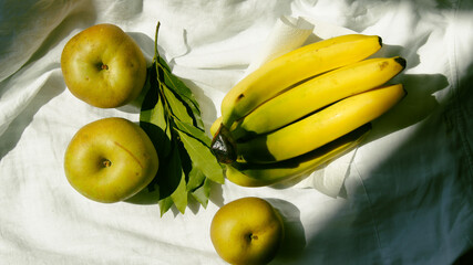 Ripe banana and green renet apples with aromatic laurel herbs, gifts of nature on white linen fabric, fruits for snacking, soft sunlight, seasonal fruits