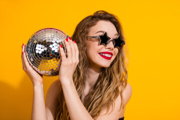 Vibrant young woman holding a disco ball, wearing star shaped sunglasses, posing joyfully in front...