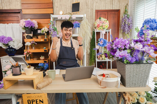 Small business flower owner shop success order first celebration happy, florist man smiling with energy at laptop, Asian store worker celebrating hands up, independent business reaching new milestone
