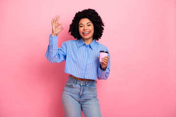 Cheerful young woman in casual outfit holding a coffee cup, signaling OK gesture with hand on pink...