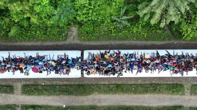 Bogura, Bangladesh - 20 August 2025: Aerial view of a train with many people riding on top of it, between lush green vegetation and parallel dirt tracks.