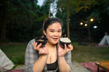 Indonesian southeast asian overweight woman holding a bowl of rice and a barbecue meal while glamping. The scene of the lifestyle of enjoying food in a natural setting