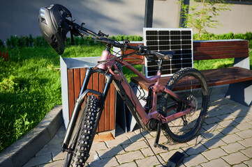 Pink electric mountain bike parked next to wooden bench, connected to solar panel for charging....